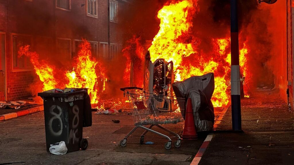 Imagen de una calle de Belfast con vehículos y viviendas dañados por el fuego